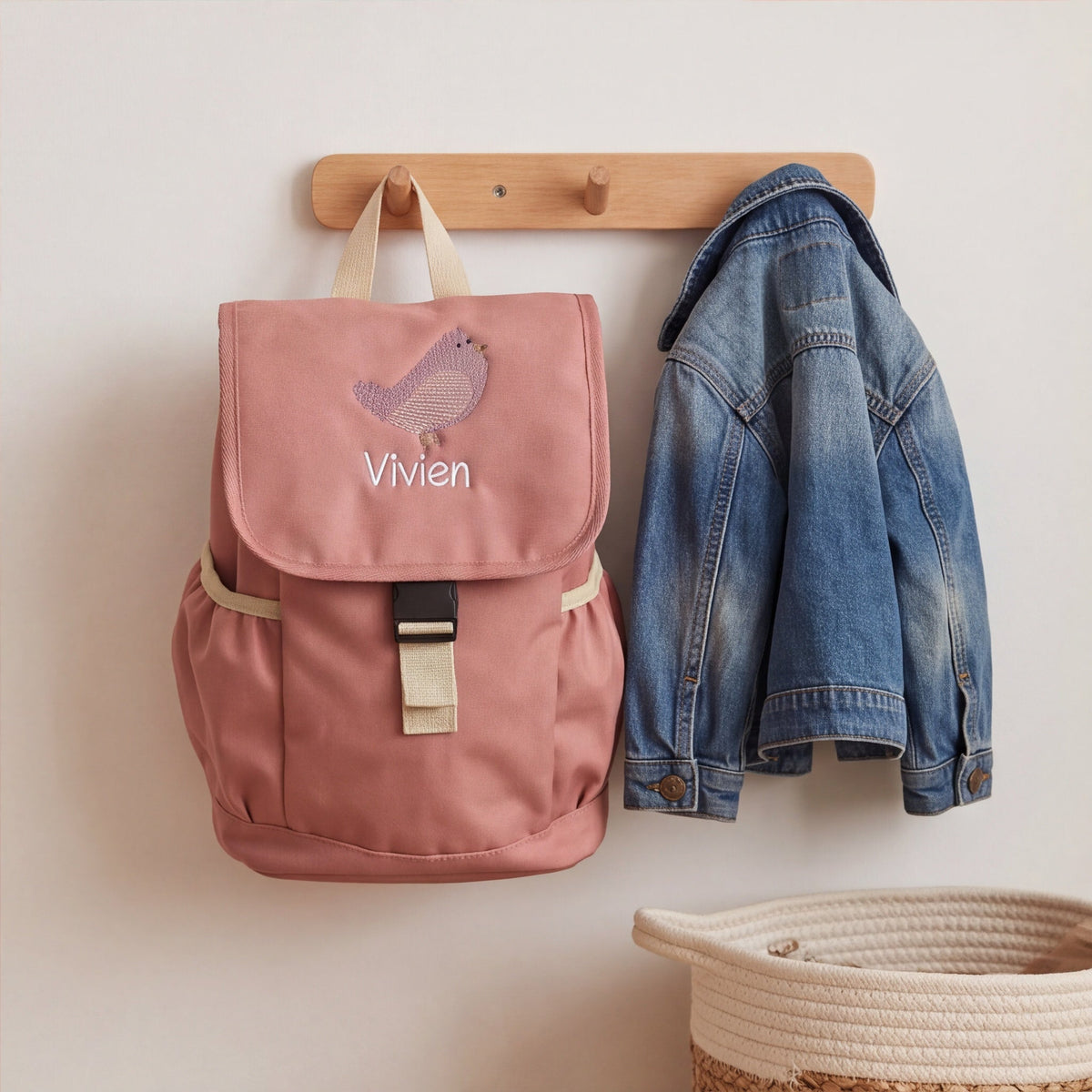 Rosa Kinderrucksack mit Vogel und Namen-Stickerei aufgehängt neben einer Jeansjacke.