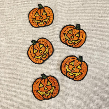Fünf orangefarbene, bestickte Kürbis-Aufnäher mit gruseligen Jack O'Lantern Gesichtern, flach angeordnet auf einem hellen Leinenstoff.