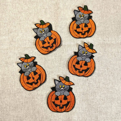Fünf bestickte Halloween-Aufnäher mit einer grauen Katze, die aus einem orangefarbenen Kürbis (Jack O'Lantern) herausschaut, angeordnet auf grauem Stoffhintergrund.