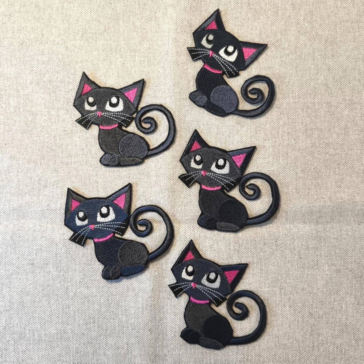 Fünf bestickte Aufnäher einer sitzenden schwarzen Katze mit großen Augen und einem auffälligen pinken Halsband, angeordnet auf grauem Stoffhintergrund.