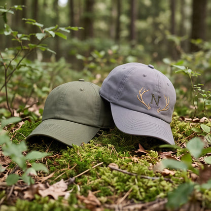 Olivengrüne und graue Dad-Hat Baseball-Caps mit Geweih-Monogramm-Stickerei nebeneinander auf Moos gebettet.