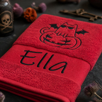 Gefaltetes rotes Handtuch mit schwarzer Namensbestickung Ella und Halloween-Katzen-Motiv neben Deko-Totenkopf