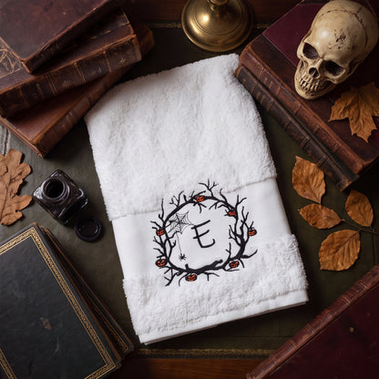 Personalisiertes weißes Handtuch mit bestickter Initiale E im Halloween Spuk-Kranz auf einem dunkelbraunen Tisch mit Herbstblätter, Bücher und einen Schädel umgeben.