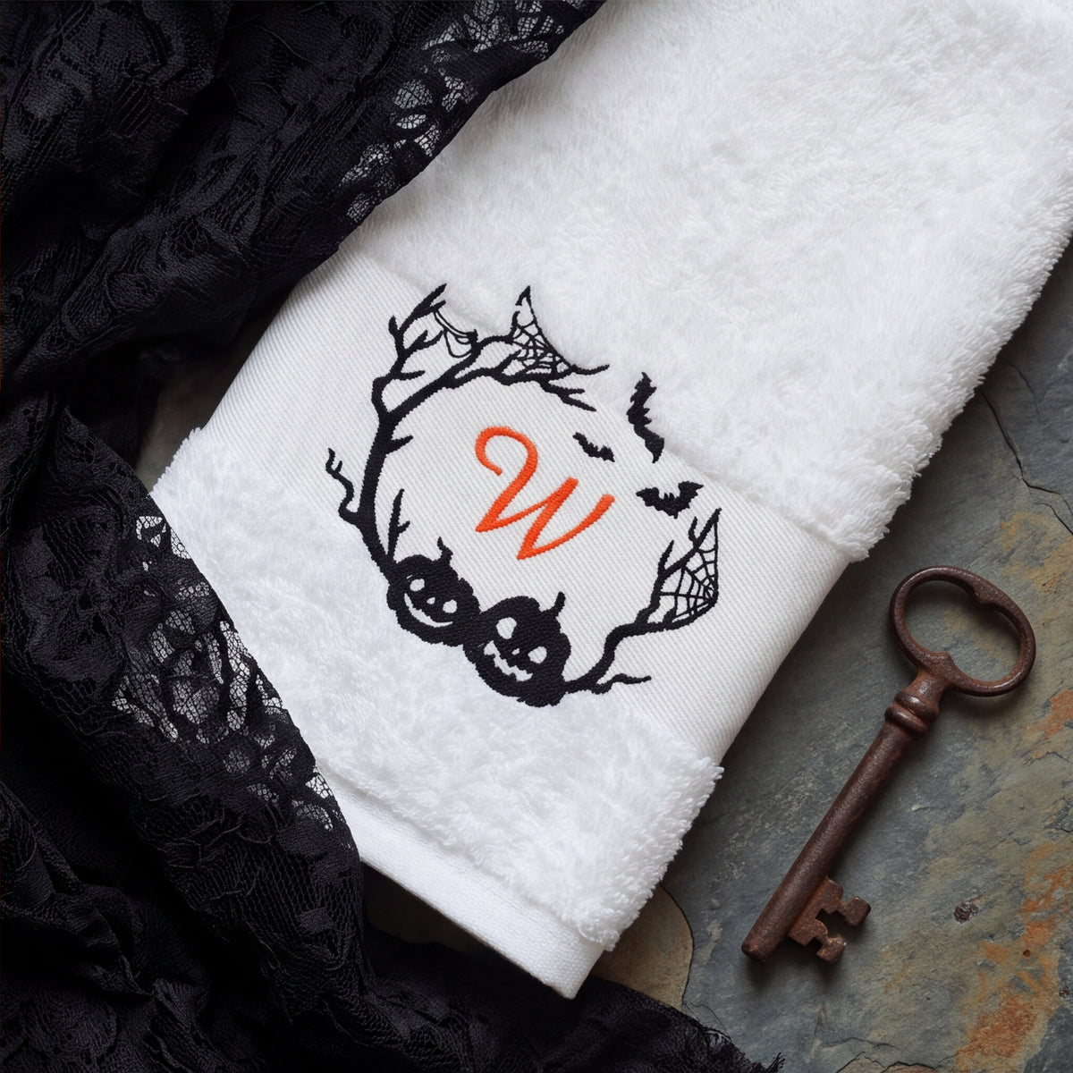 Detailaufnahme des bestickten Halloween-Monogramms mit Fledermäusen und Kürbissen auf weißem Frottier neben schwarzer Spitze