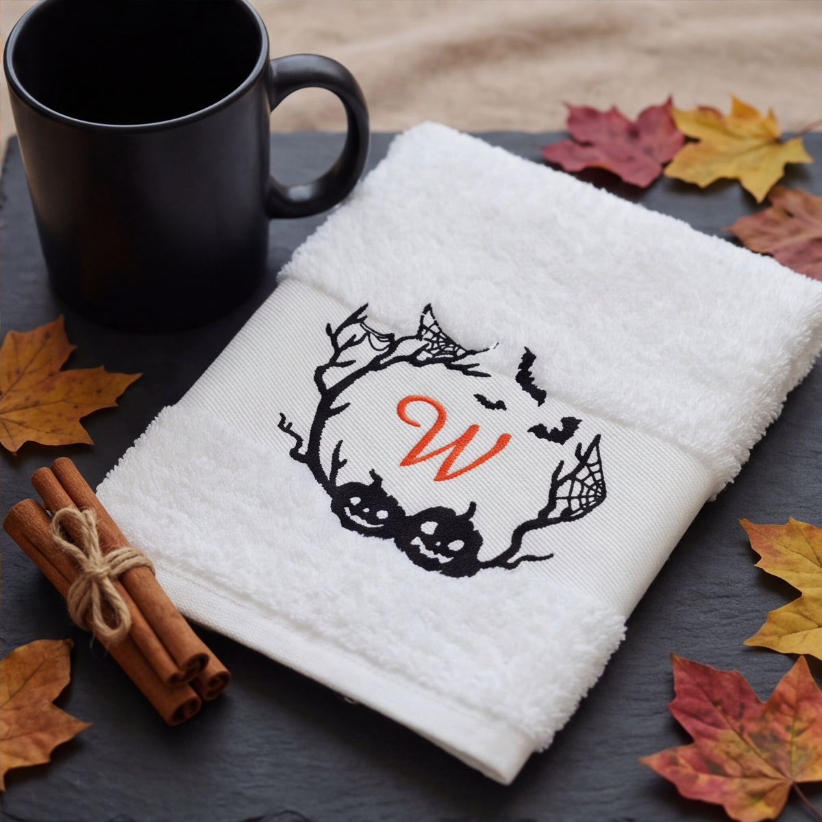 Zusammengelegtes weißes Handtuch mit orange besticktem W und Halloween-Spuk-Kranz neben Kaffeetasse und Herbstblättern