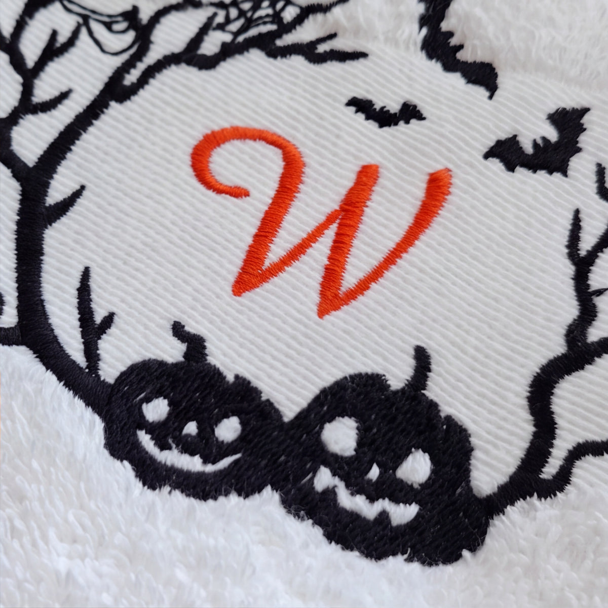 Makroaufnahme des orange gestickten Buchstaben W und der schwarzen Kürbis-Bestickung auf weißem Baumwolltuch