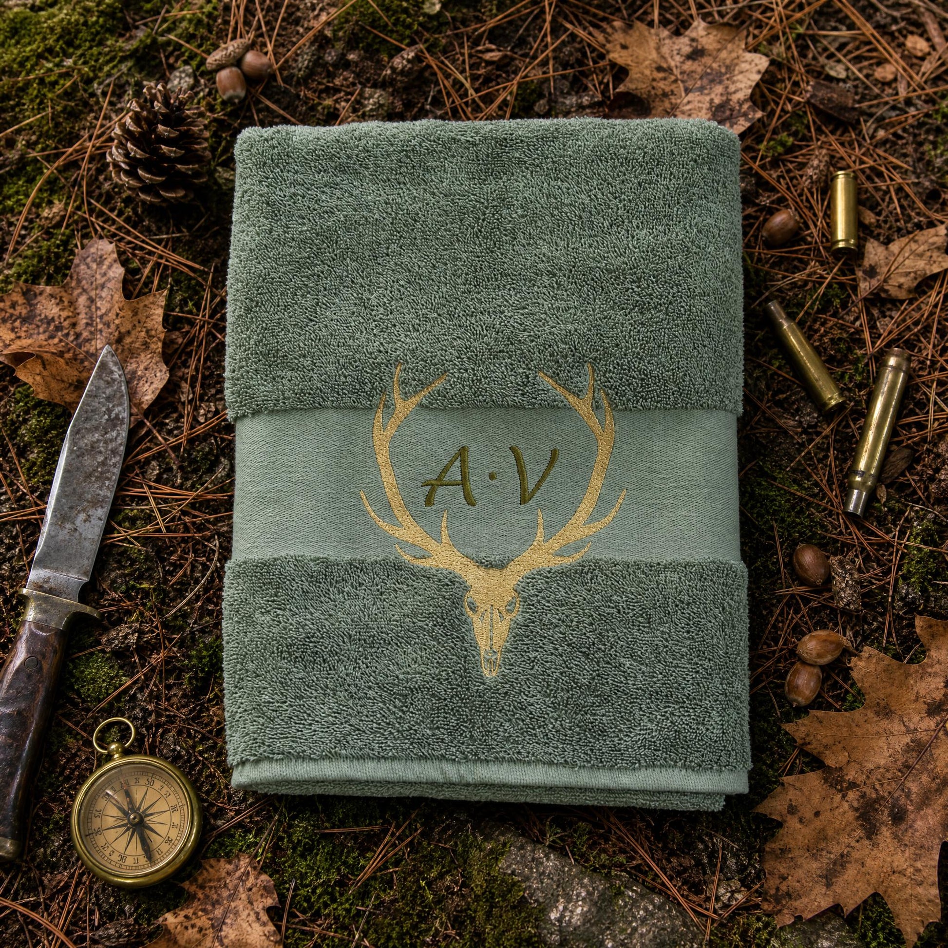 Olivgrünes handtuch mit goldener Hirschschädel und Geweih sowie Initialen Bestickung auf einem natürliche Waldboden Hintergrund.