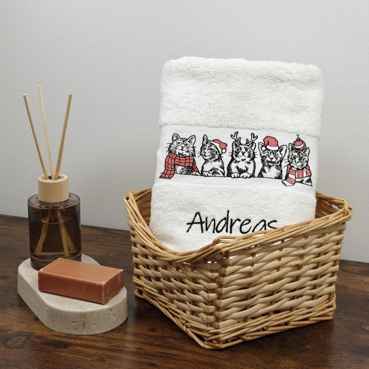 Weißes flauschiges Handtuch in einem Weidenkorb, bestickt mit fünf Katzen-Illustrationen mit roten Nikolausmützen und Schals sowie dem Namen Andreas in schwarzer Schreibschrift.