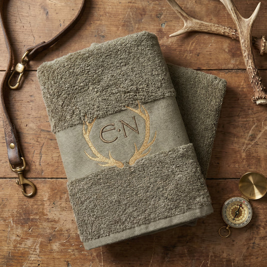 Zwei olivgrüne Handtucher auf dunkelbraunem holz umgeben von Jägerausrüstung, mit einer goldenen Hirschgeweih und khaki Initialen Bestickung