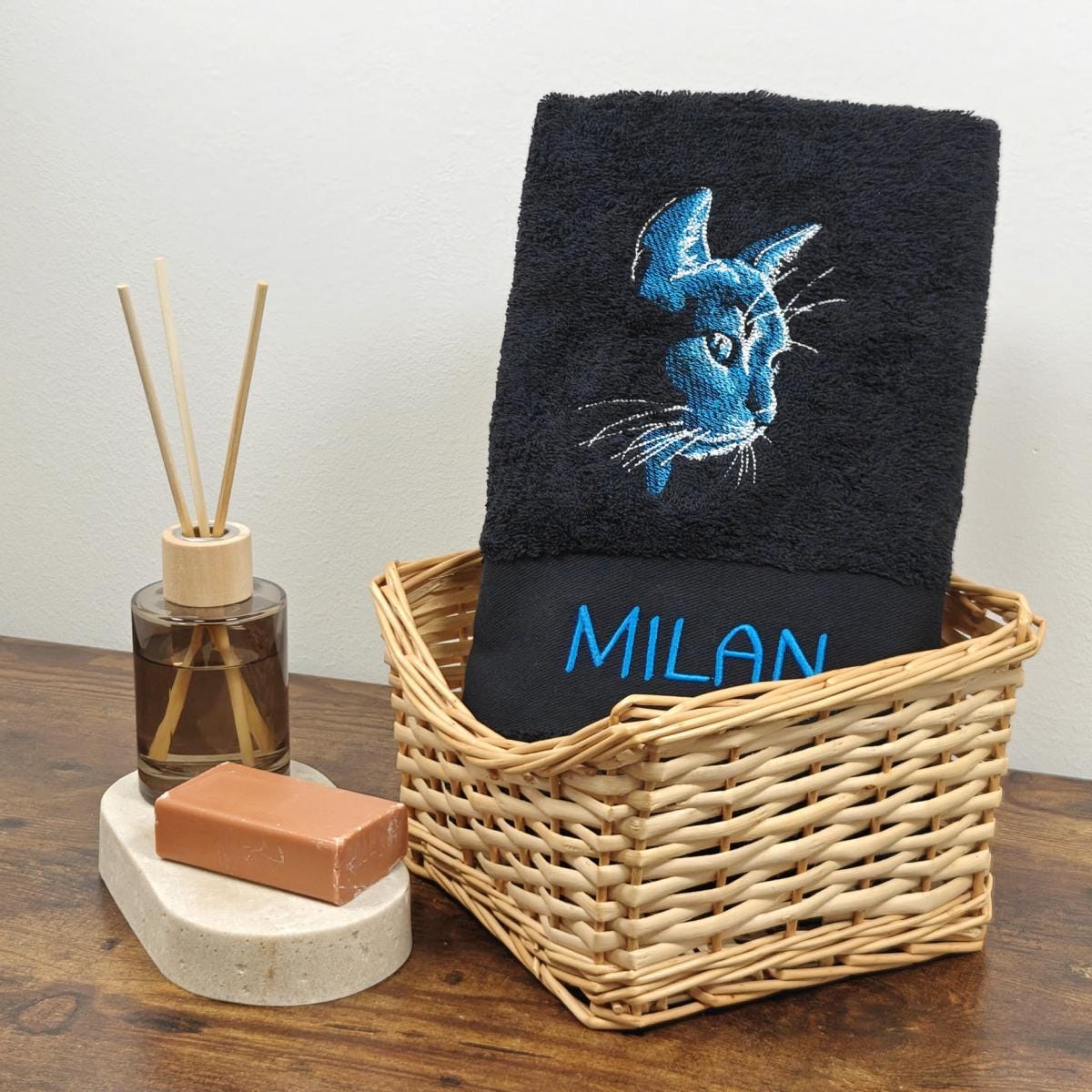 Schwarzes besticktes Handtuch mit blauem Katzenkopf-Profil und dem Namen Milan in leuchtend blauem Garn, präsentiert in einem Weidenkorb mit Bad-Accessoires.