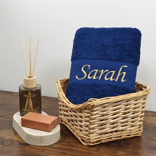 Ein blaues Handtuch mit Namen in Gold bestickt in einem hellen Korb auf einem dunkelbraunen Tisch neben einer Seife und Duftdiffusor aufgestellt