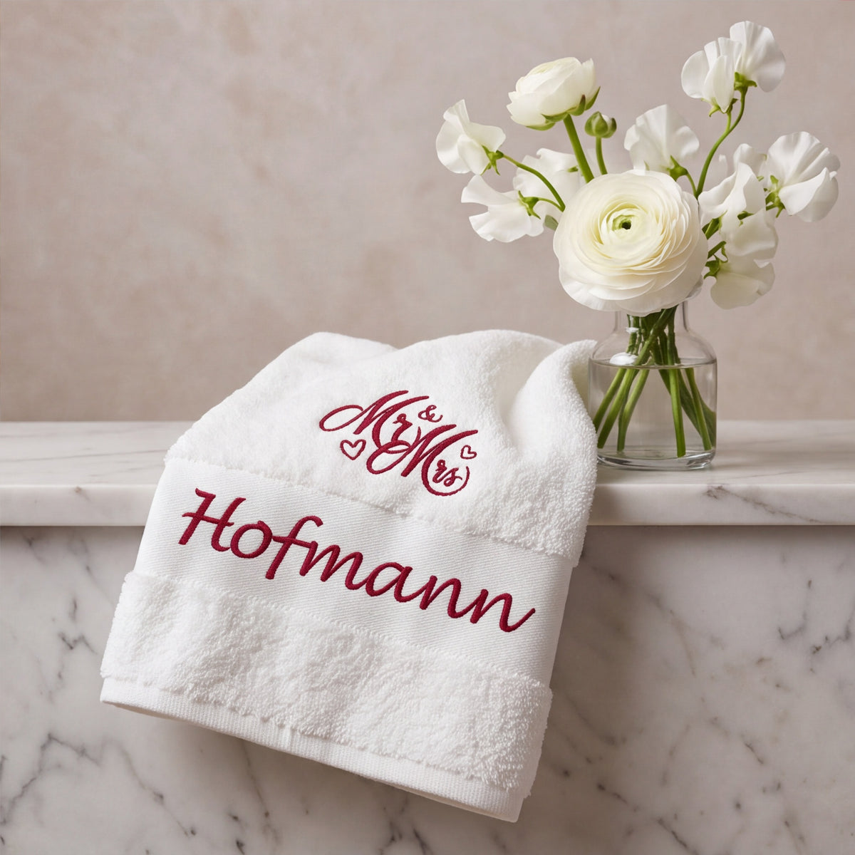 Elegantes weißes Handtuch mit weinroter Namensbestickung Hofmann auf Marmorablage neben weißer Blumenvase