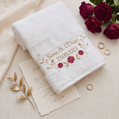 Besticktes Hochzeits-Handtuch in Weiß mit edlem Rosen-Ranken-Motiv und Wunschtext auf einer Einladungskarte neben Eheringen und roten Rosen.