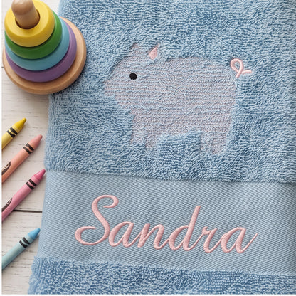 Nahaufnahme der rosa Namensstickerei Sandra und der Schweinchen-Kontur auf flauschigem blauen Frotteestoff neben Holz-Stapelringen und Wachsmalstiften.