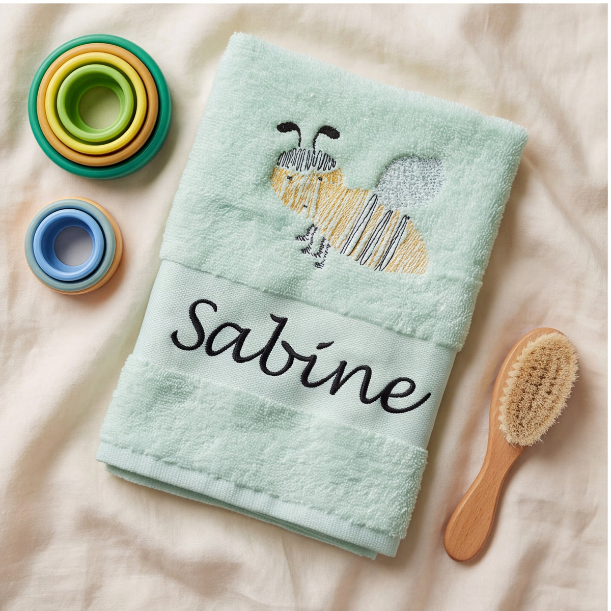 Besticktes mintfarbenes Babytuch mit Biene und Namenszug Sabine neben Stapelbechern und einer Holz-Babybürste.