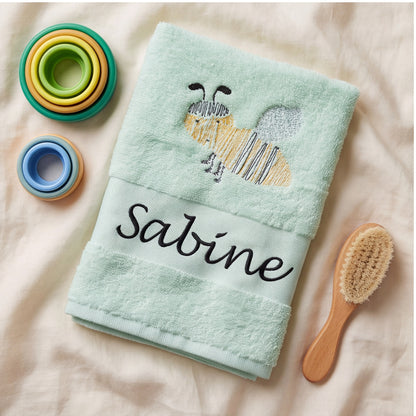 Besticktes mintfarbenes Babytuch mit Biene und Namenszug Sabine neben Stapelbechern und einer Holz-Babybürste.