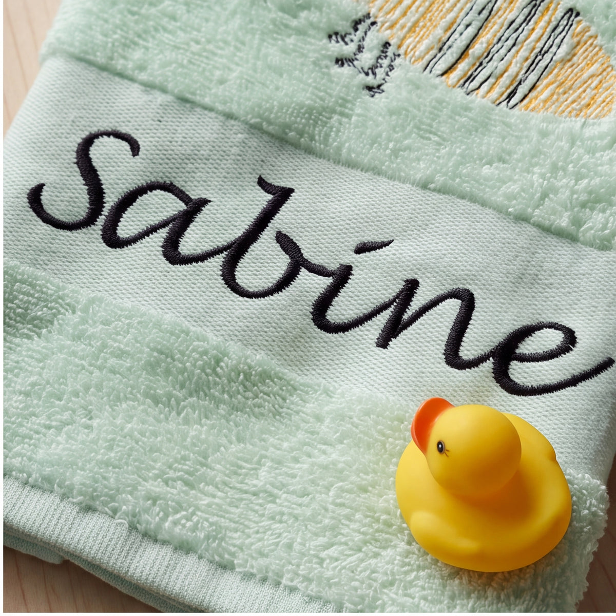 Nahaufnahme der dunklen Namensstickerei Sabine auf dem mintgrünen Handtuch mit einer gelben Badeente im Vordergrund.