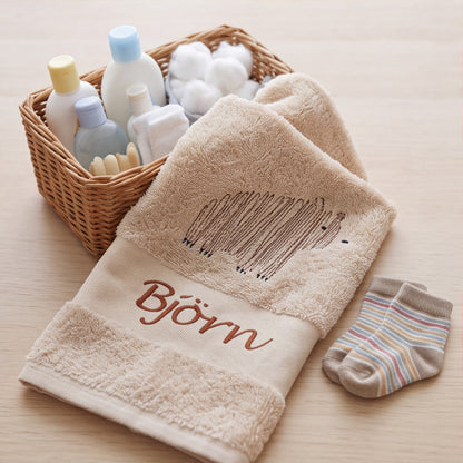 Besticktes Handtuch für Kinder in Beige mit Bärenmotiv und dem Namen Björn neben einem Pflegekörbchen und Babysöckchen.