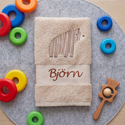 Beige zusammengelegtes Kinderhandtuch mit Bären-Stickerei und Namenszug Björn, umgeben von bunten Stapelringen auf einer grauen Filzunterlage.