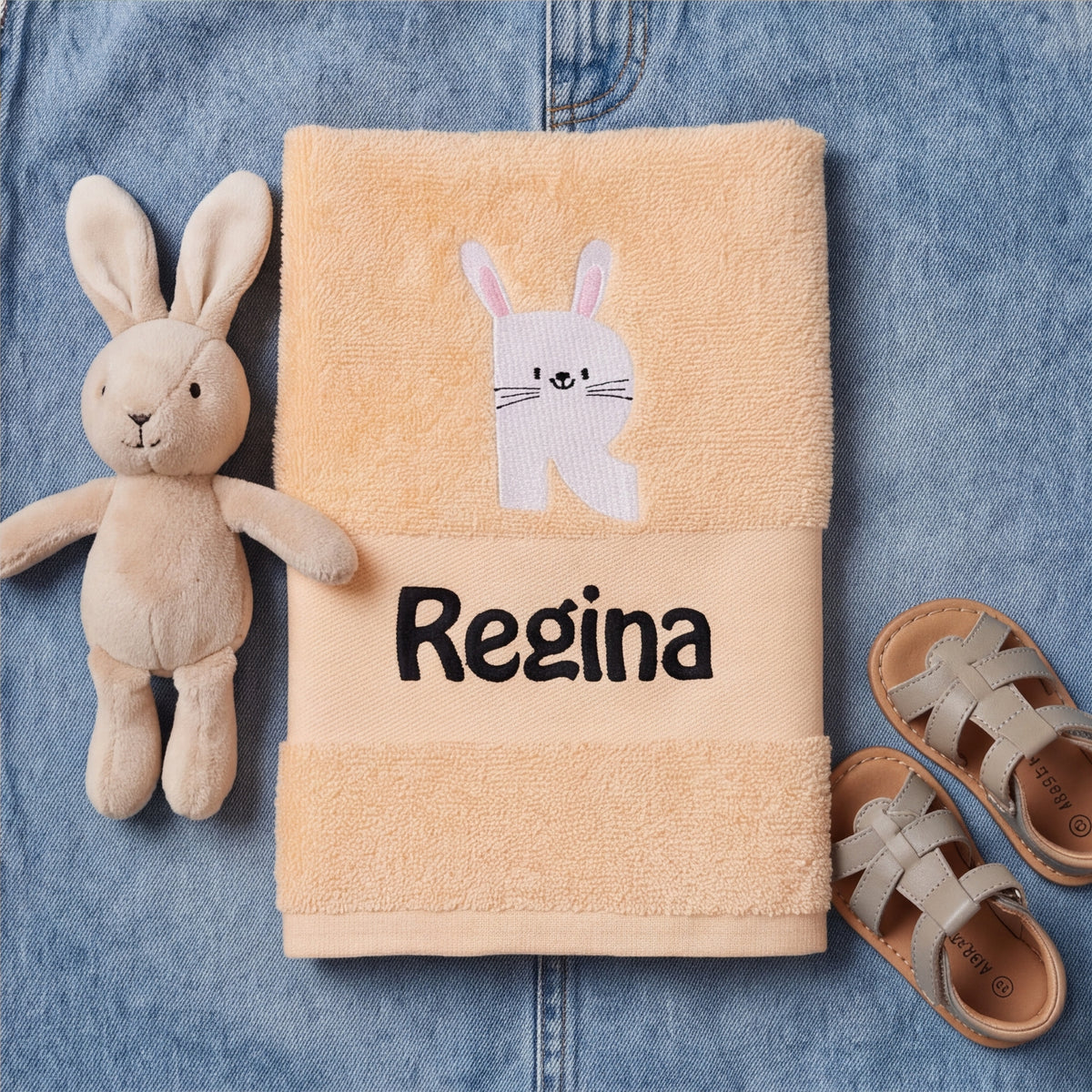 Pfirsichfarbenes Kinderhandtuch mit gesticktem Hasen-Initial R und Namenszug Regina auf Jeansstoff neben einem Kuschelhasen und Kindersandalen.