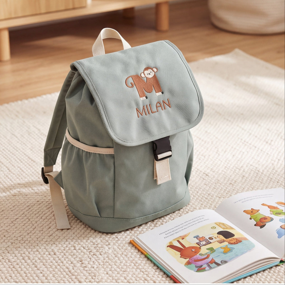 Graugrüner Kinderrucksack Dusty Green mit besticktem Affen-Buchstaben M und dem Namen Milan neben einem aufgeschlagenen Kinderbuch.