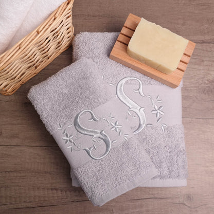 Zwei graue Handtücher mit gesticktem silbernen Buchstaben "S" und Blumendesigns. Die Handtücher sind gefaltet und auf einer Holzoberfläche gestapelt.