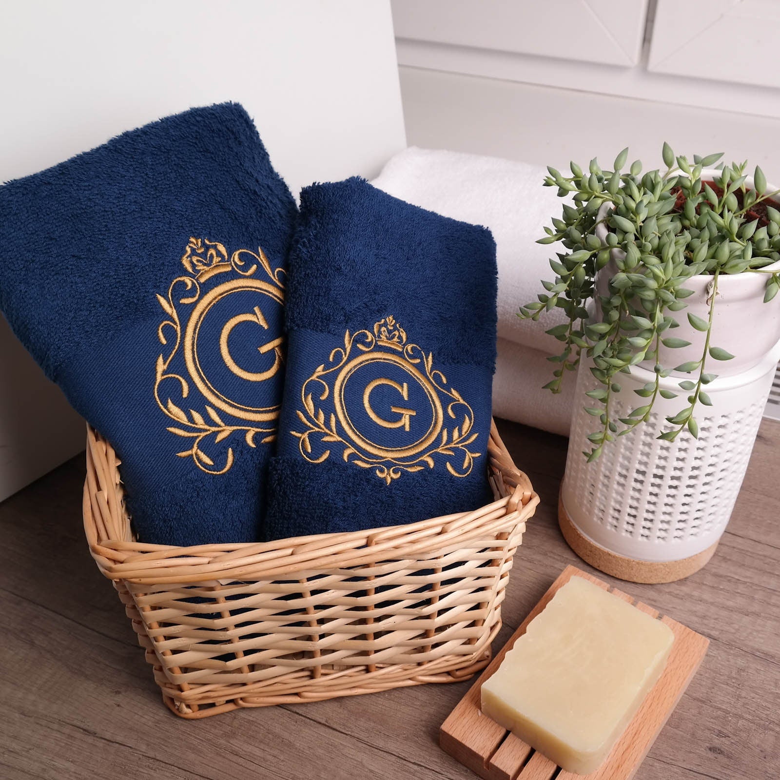 Zwei dunkelblaue, flauschige Handtücher in einem Weidenkorb, bestickt mit einem goldenen Royal-Wappen und dem Buchstaben G, daneben eine Pflanze und Naturseife auf Holz.