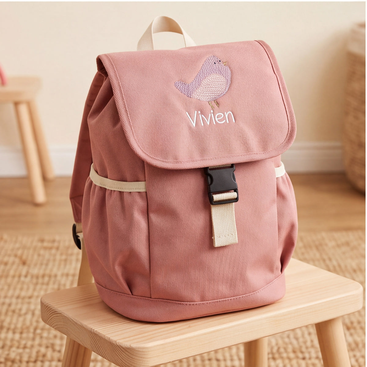 Personalisierter rosa Krippenrucksack mit Namensstickerei Vivien und lila Vogel auf einem Holzstuhl im Kinderzimmer.