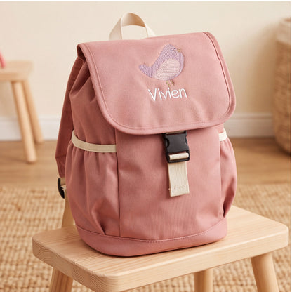 Personalisierter rosa Krippenrucksack mit Namensstickerei Vivien und lila Vogel auf einem Holzstuhl im Kinderzimmer.