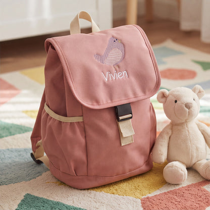 Altrosa Kinderrucksack mit besticktem Vogel-Motiv und dem Namen Vivien, stehend auf einem Spielteppich neben einem Teddybären.
