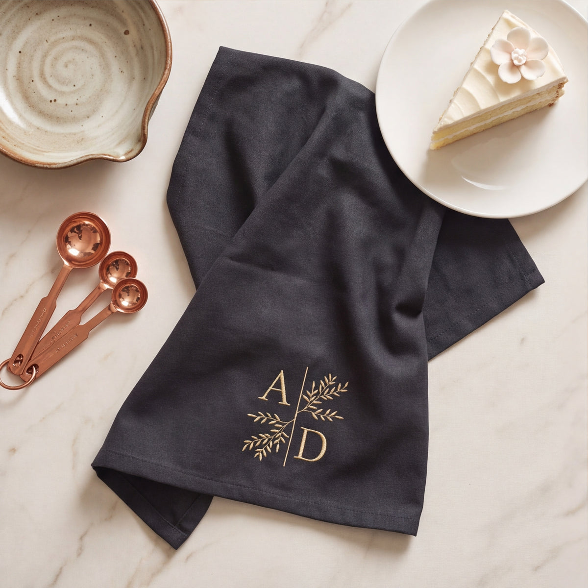 Schwarzes Bio-Geschirrtuch locker drapiert auf Marmor mit gold gesticktem Zweig-Monogramm A D, Hochzeitstorte und Messlöffeln