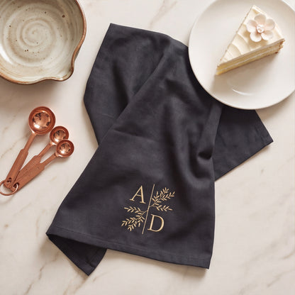 Schwarzes Bio-Geschirrtuch locker drapiert auf Marmor mit gold gesticktem Zweig-Monogramm A D, Hochzeitstorte und Messlöffeln