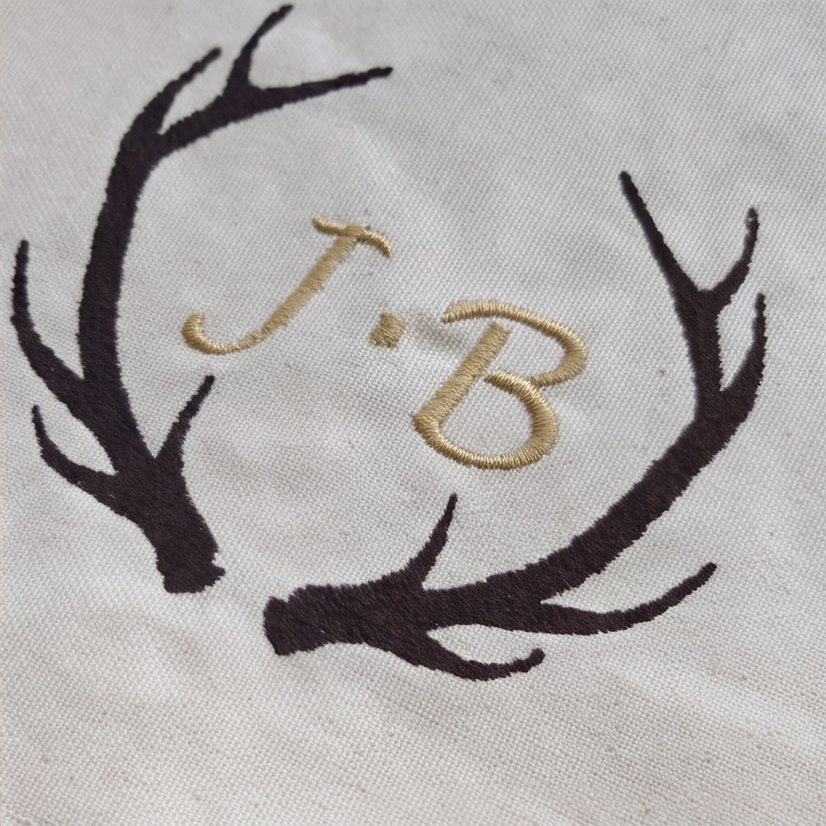 Nahaufnahme der braunen Geweih-Stickerei mit gold gestickten Initialen J.B. auf naturfarbenem Bio-Baumwollstoff