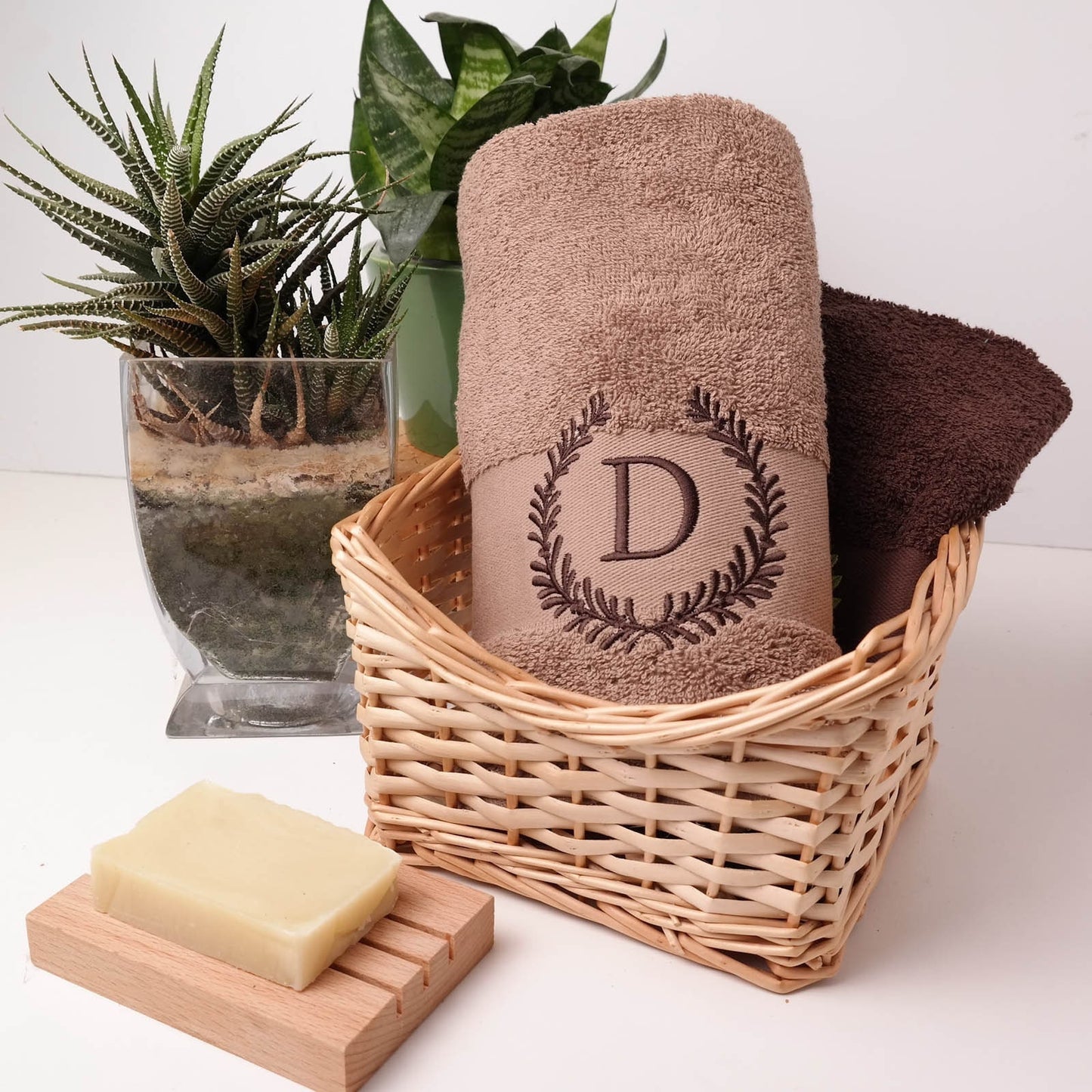 Sandfarbenes (Taupe) flauschiges Handtuch in einem Weidenkorb, bestickt mit einem dunkelbraunen Blätterkranz und dem Initial D, harmonisch dekoriert mit Zimmerpflanzen und Naturseife.