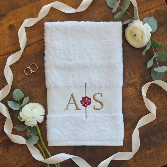 EIn weißes personalisiertes Handtuch mit goldenen Initialen A und S, sowie einer Linie mit einer Rose mittig bestickt. Das Handtuch ist umgeben von Hochzeitsdekorationen auf einem dunkelbraunen Tisch