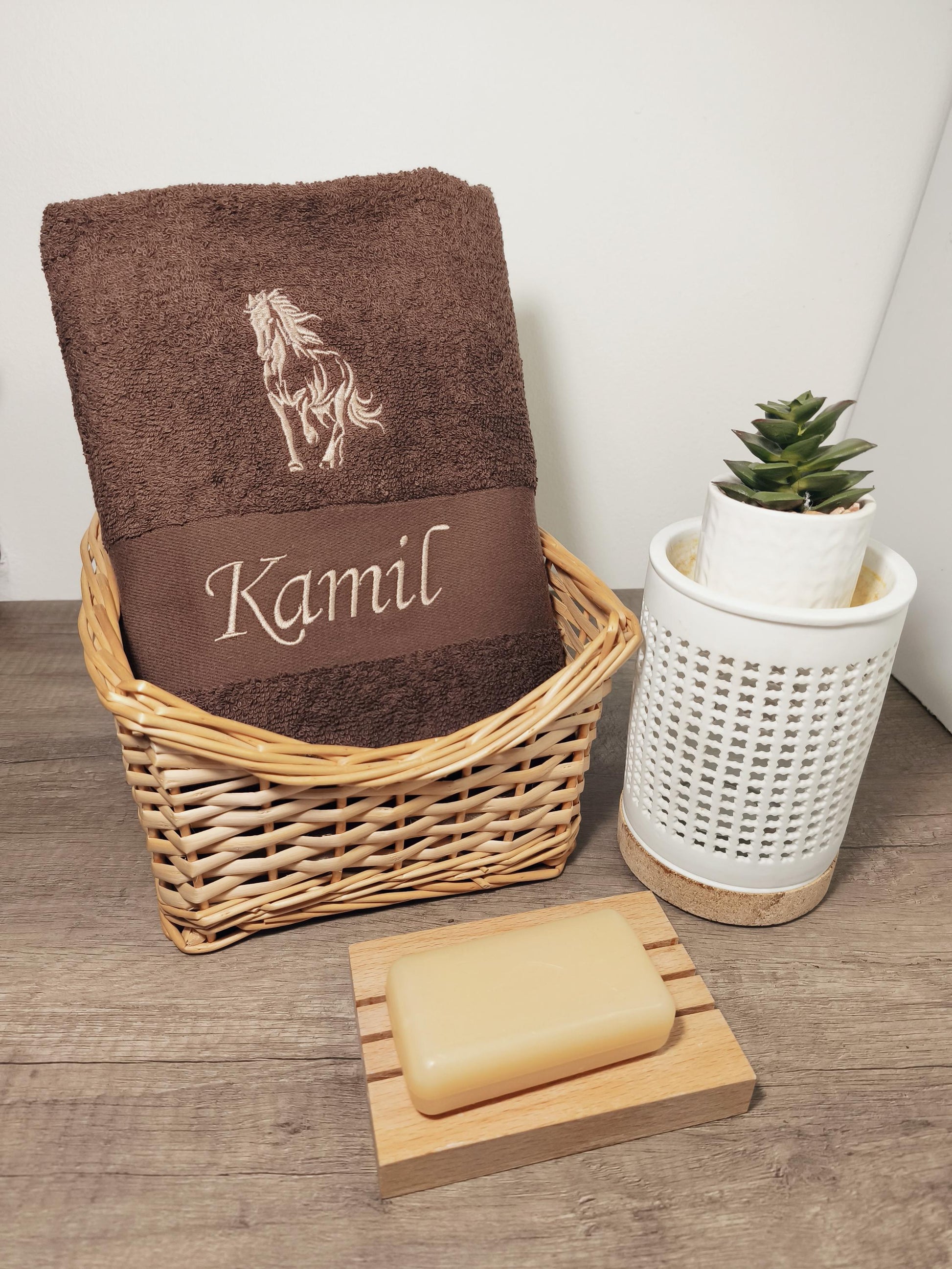 Dunkelbraunes, flauschiges Handtuch in einem Flechtkorb, bestickt mit einem galoppierenden Pferd und dem Namen Kamil in heller Garnfarbe, dekoriert mit Seife und einer kleinen Pflanze.