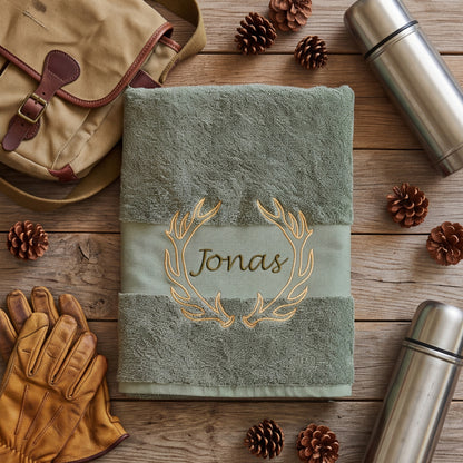 Olives Handtuch mit gesticktem Geweih-Motiv und dem Namen Jonas flach zusammengelegt auf Holz neben Ledertasche, Thermosflaschen und Tannenzapfen.